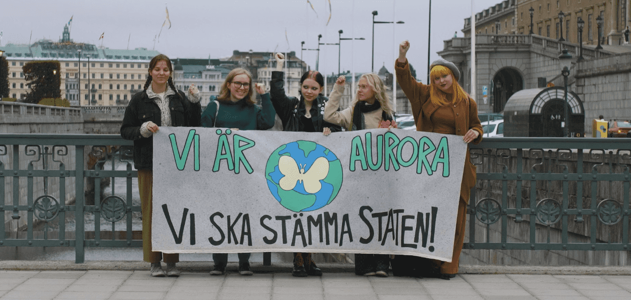 Five young people standing with a banner outside the Parlament building in Stockholm. The banner reads: We are Aurora, We will sue the state!